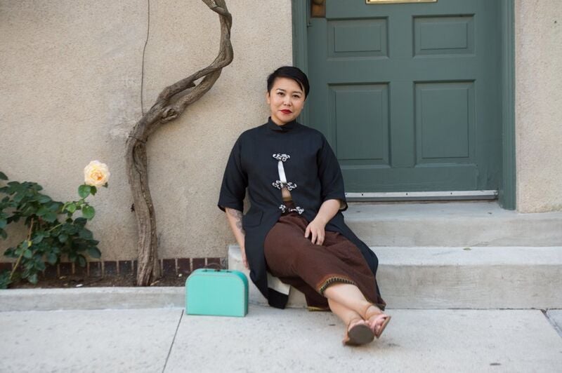 A woman in a black top and brown skirt sits on a sidewalk step in front of a green door, a turquoise bag beside her.