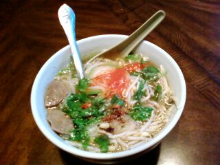 A white bowl of noodle soup with sliced meat, a red garnish, green herbs and bean sprouts, two spoons in it.