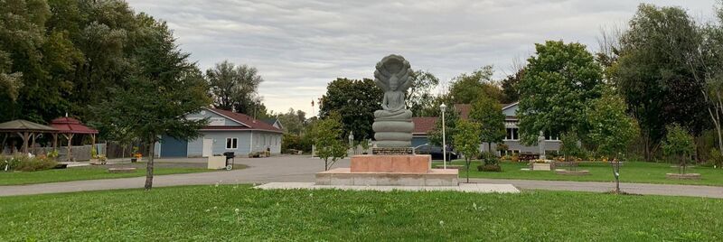 Lawn and driveway with a grey stone Buddha seated under a seven-headed naga on a pink plinth, blue-sided temple buildings behind.
