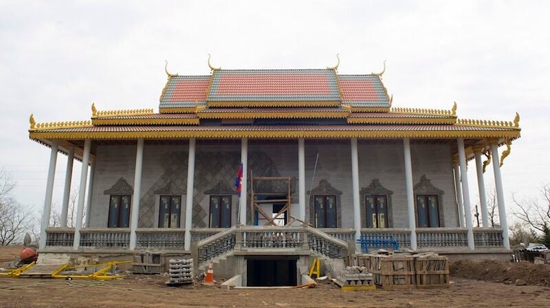 Large Khmer-style temple hall with grey walls, golden roof edges and front stairs, surrounded by bare ground and building materials.