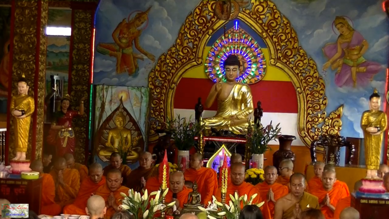 Monks in saffron robes seated before a shrine with a gilded Buddha and painted murals.