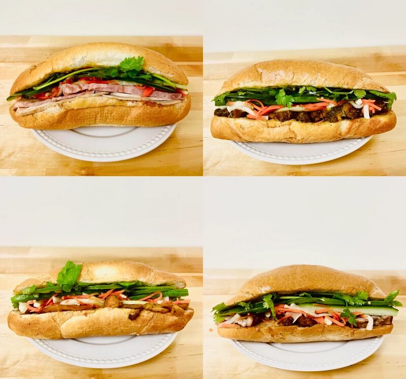 Four baguette sandwiches filled with meat, pickled carrot, cucumber and herbs, each on a white plate on a wooden counter.