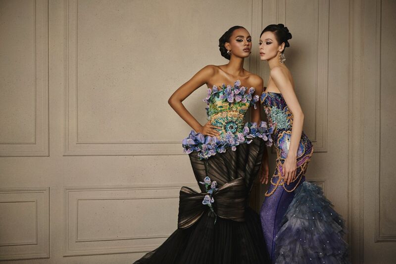 Two models in sculpted strapless gowns with blue and green floral bodices pose against a panelled wall.