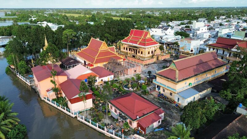An aerial view of a riverside temple compound with red-roofed Khmer halls, smaller buildings and gardens among trees.