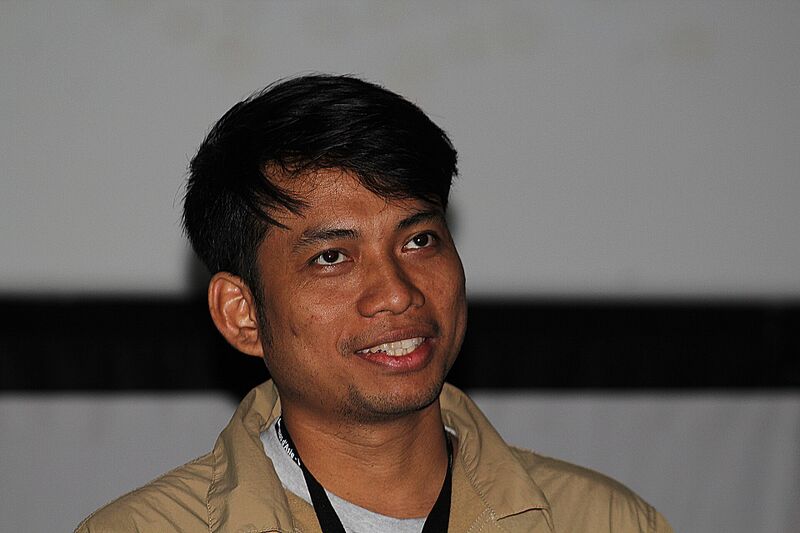 A smiling man with short black hair in a beige jacket and lanyard.