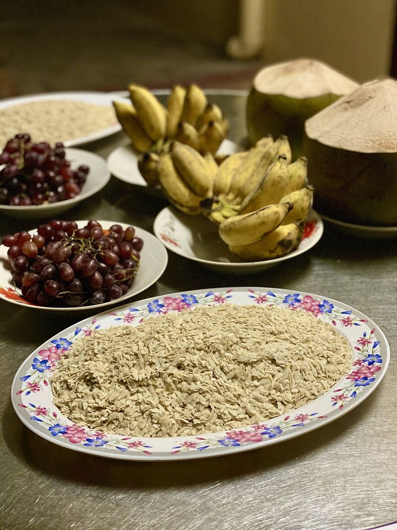 A decorated plate of pale flattened rice beside bananas, coconut and bowls of red grapes on a wooden table.