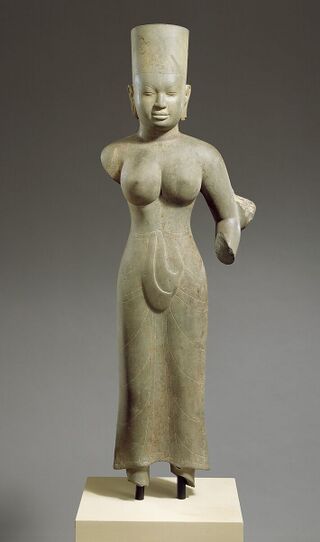 A stone statue of a standing woman in a tall headdress, wearing a long skirt cloth that falls to the ankles.