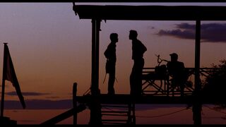 Silhouettes of three men on a wooden watchtower against a purple evening sky, with a flag at the left.