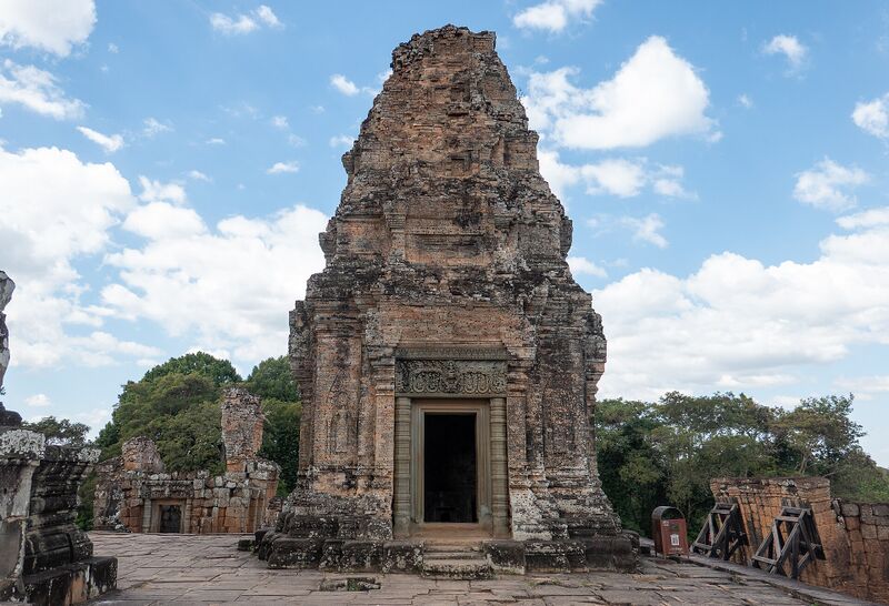 A tall laterite and brick temple tower with a doorway, standing on a stone terrace under a blue sky with white clouds.