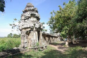 A weathered sandstone tower with carved panels stands at the end of a long stone wall. A dirt path runs beside the wall under leafy trees, with tall grass and a blue sky on the left.
