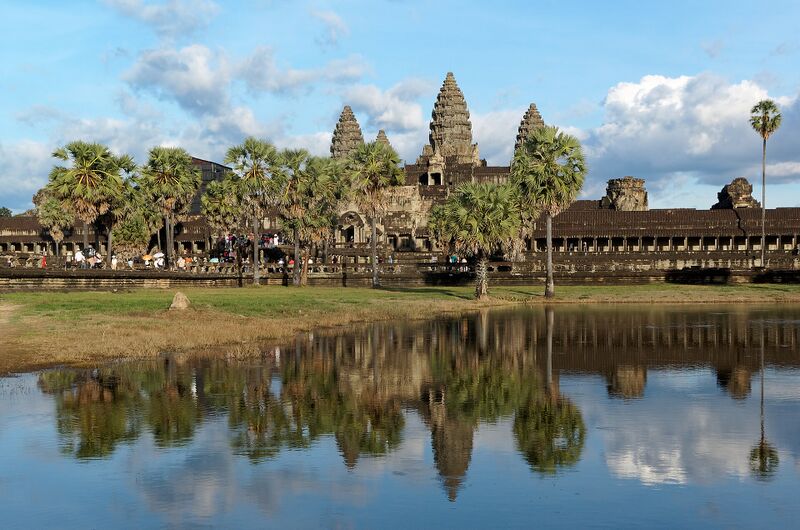 Five sandstone towers of Angkor Wat rising behind palm trees, mirrored in a still pool under a blue sky.