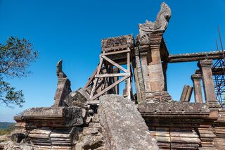Wooden scaffolding propping up leaning sandstone walls and a door frame on a rocky platform.