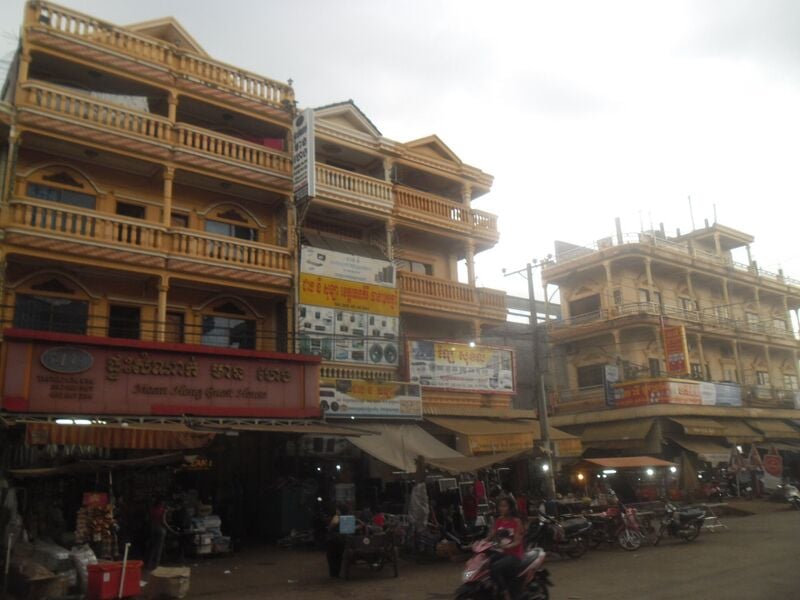 Multi-storey shophouses with balconies and shop signs along a dusty street in Banlung, with motorbikes parked in front