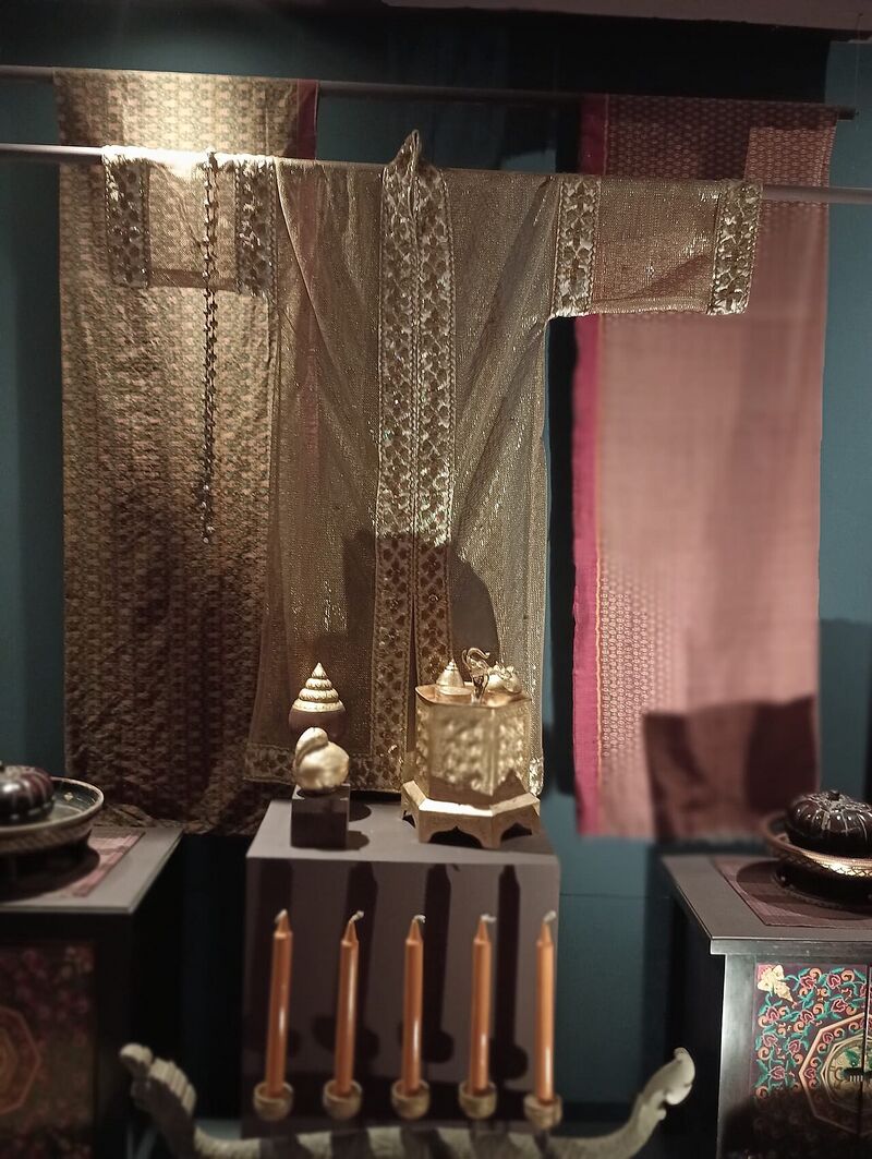 Sheer gold-brown robes with long sleeves and wide patterned borders hung against a wall, above a table with offerings and candles.
