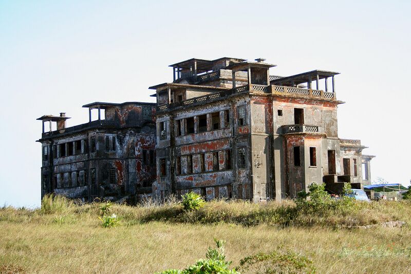 A weathered, multi-storey hotel with flat roofs and blackened walls standing alone on a grassy plateau under a pale sky.