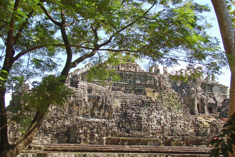A broad stepped stone temple seen through the branches of a large tree, its long western side built up in tiers.