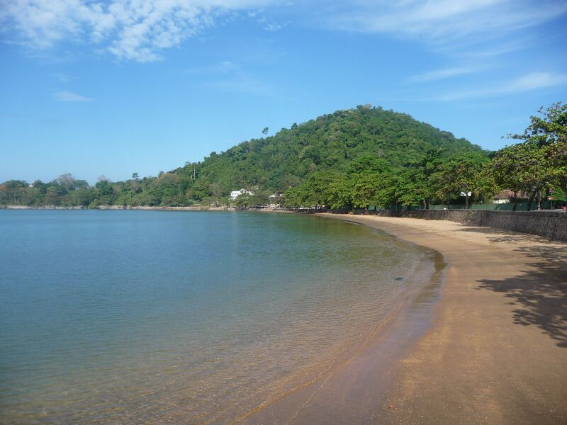 Calm, clear water laps a curving strip of golden sand that runs along a low stone seawall shaded by broad trees. A rounded hill covered in dense green forest rises behind the bay under a blue sky with thin clouds, with a few white buildings at its foot.