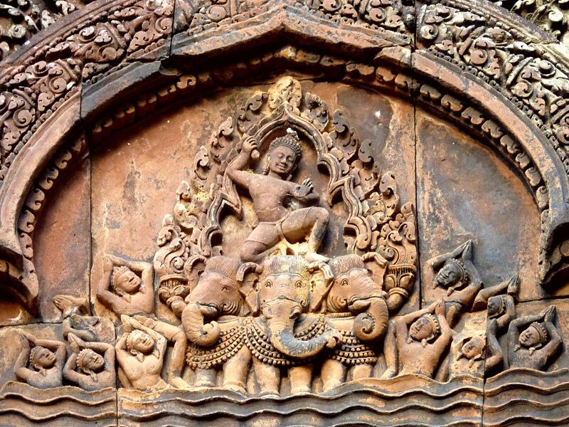 A carved pink sandstone pediment in which a crowned god, one arm raised, crouches on the heads of a three-headed elephant, small figures at its feet, under a curved, richly carved frame.
