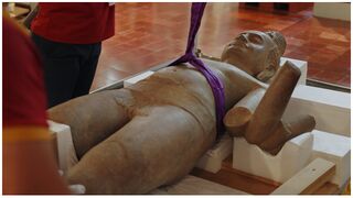 A headless sandstone statue lies in a padded wooden crate on a polished floor while people stand nearby.
