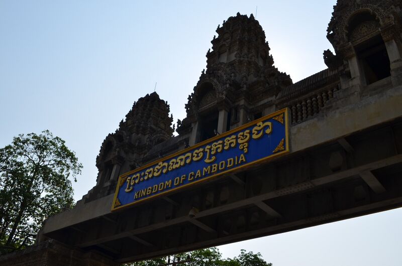 Three carved stone towers in the style of an Angkor temple rise above a gateway beam that carries a blue and gold sign in Khmer and English reading Kingdom of Cambodia, seen from below against a pale sky, with a tree at the left.