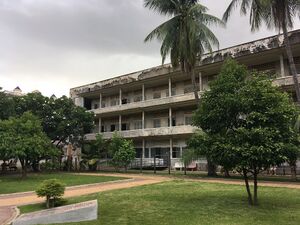 A long three-story concrete building with open walkways and shuttered windows, seen across a lawn with young trees and palms under a grey sky.