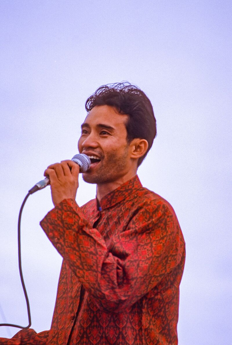 Arn Chorn-Pond, in a red patterned shirt, sings into a microphone outdoors against a pale sky.