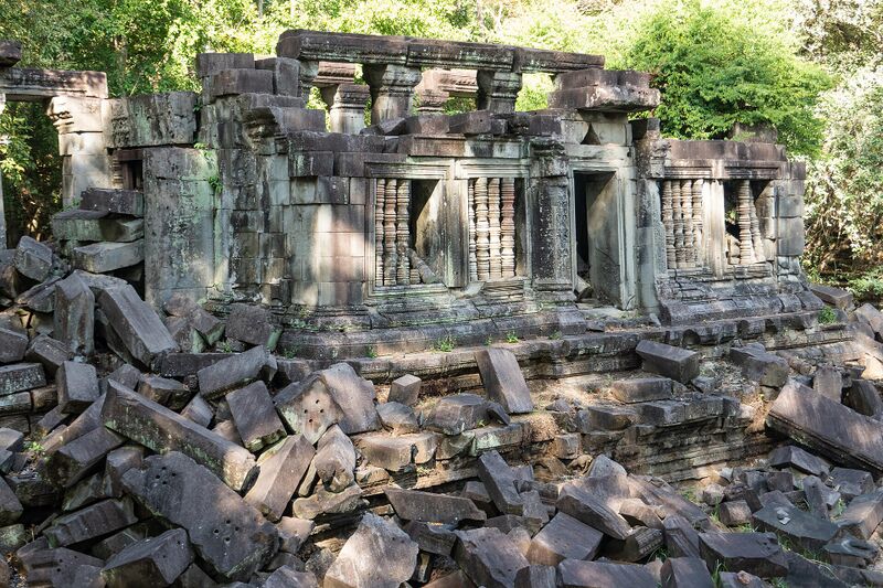 A ruined stone gallery with barred windows behind a slope of tumbled sandstone blocks and trees.