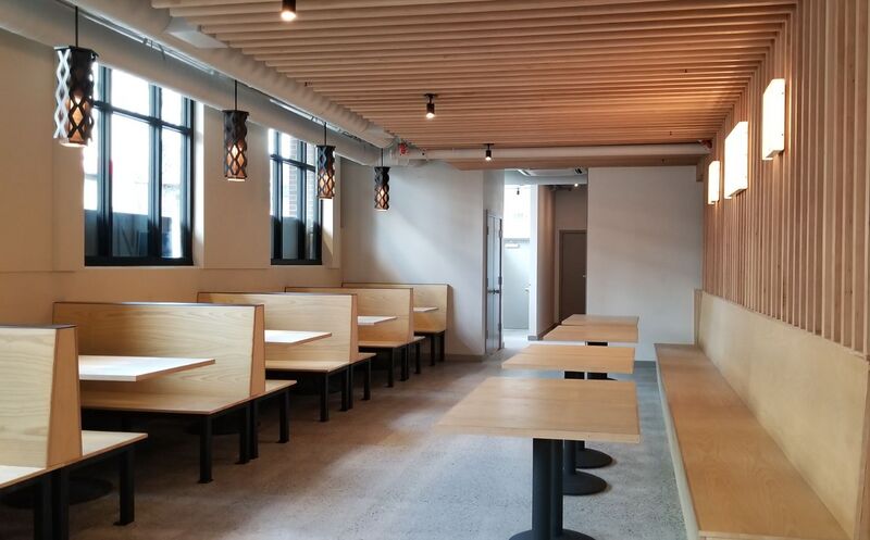 A bright restaurant dining room with wooden booths, tables and pendant lights along a window wall.