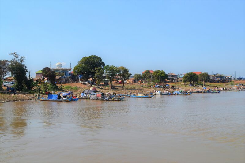 Wooden fishing boats, many under blue and gray tarps, line the muddy bank of a wide brown river. Behind them are trees, houses on stilts and, at the left, a white building with a round dome, under a clear blue sky.