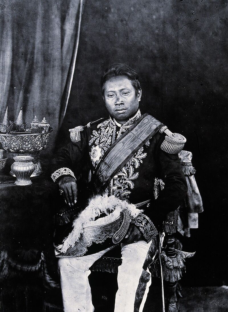 Black-and-white studio portrait of a seated man in an embroidered uniform with epaulettes and a sash, a plumed hat on his lap and ornate silver vessels on a table beside him.