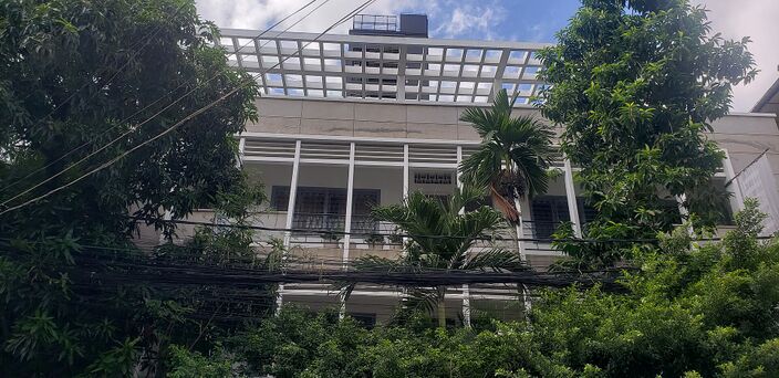 The upper floors of a white building with balconies and louvred windows, seen above a screen of green trees.
