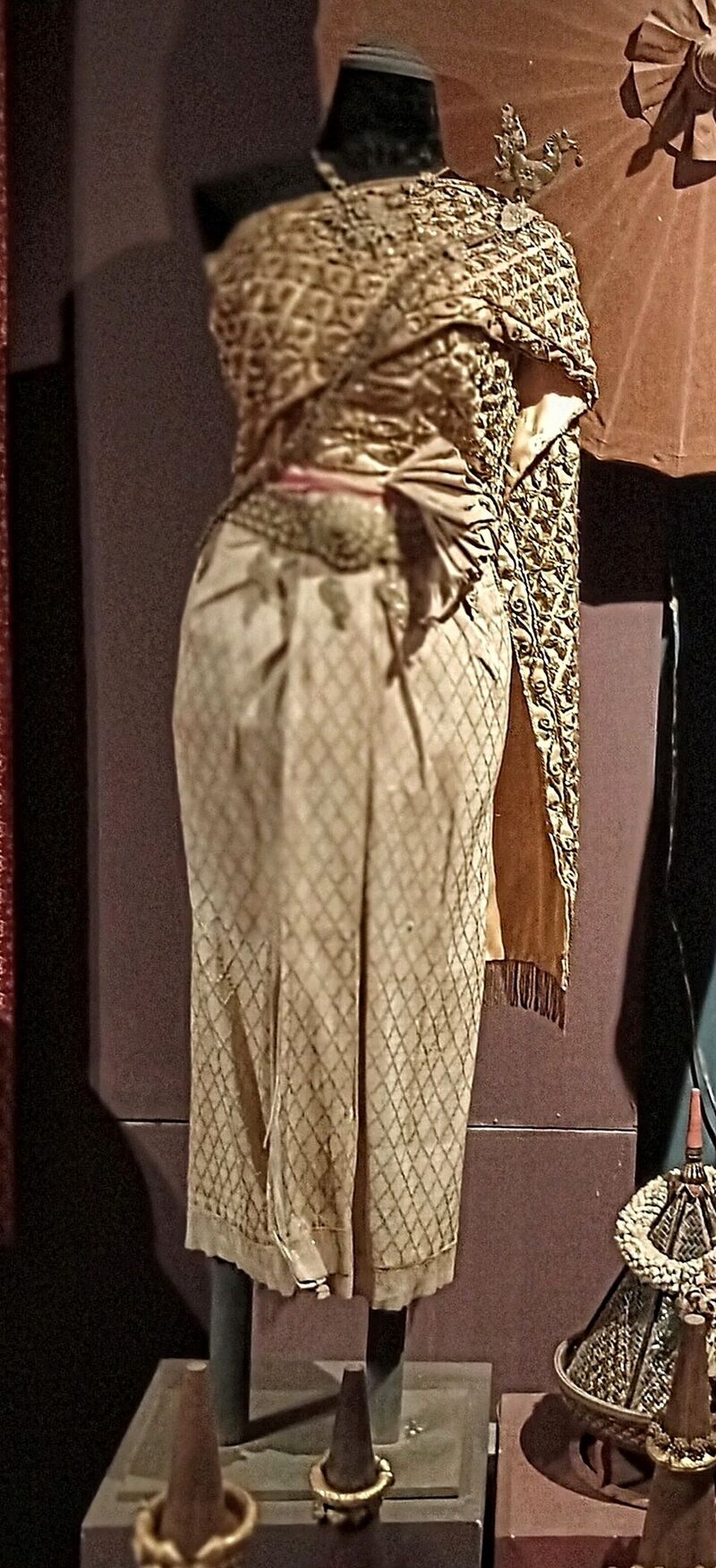 A cream and gold brocaded sampot and a matching shawl on a dress stand against a dark wall.