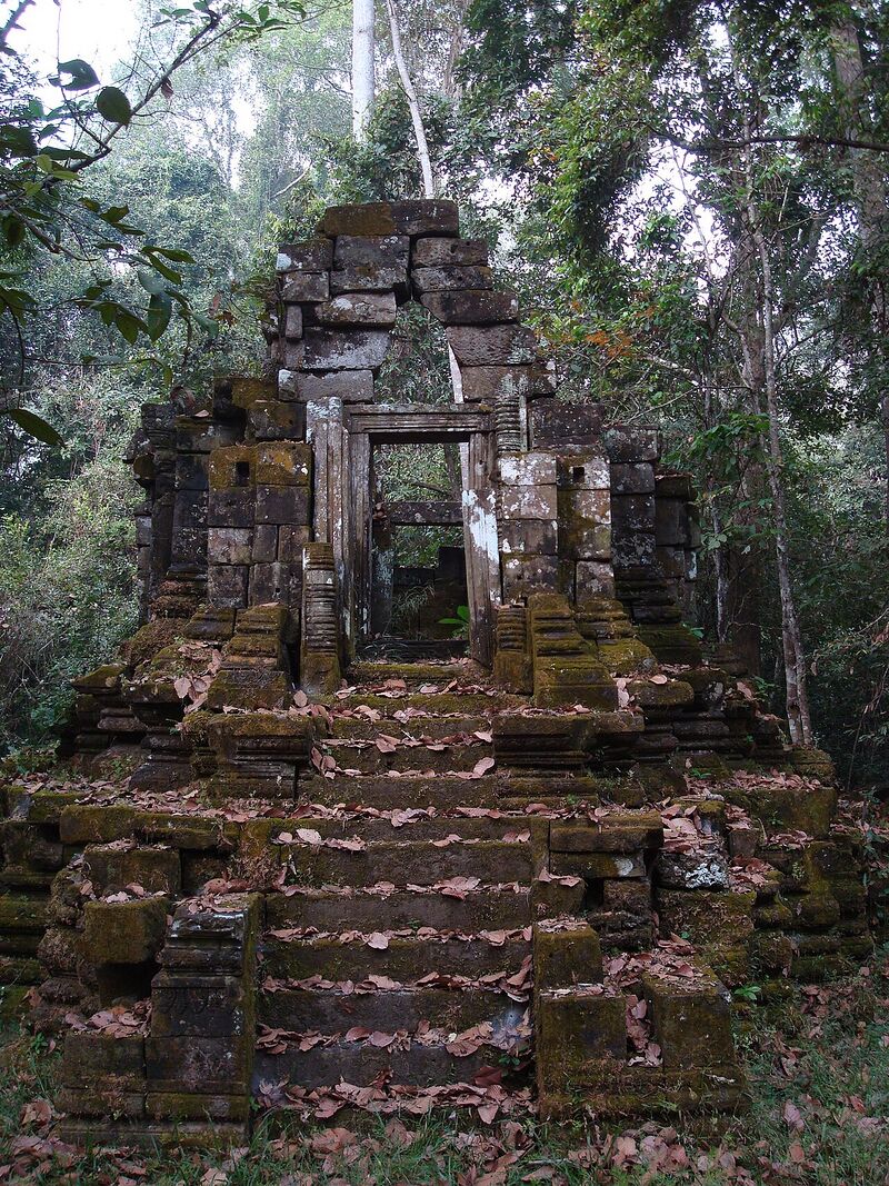 A small moss-covered sandstone sanctuary with an open doorway and a corbelled gable, at the top of a steep flight of leaf-strewn steps in the forest.