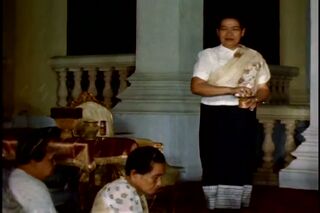 A woman in a white blouse and dark sampot stands between palace pillars, holding an offering, with seated women nearby.