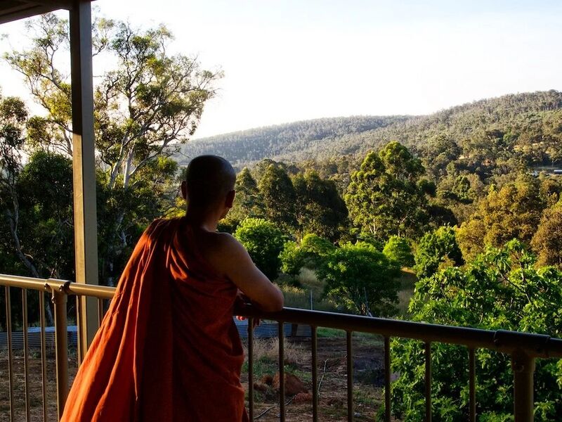 A monk in an orange robe, seen from behind, leans on a veranda railing above forested hills.