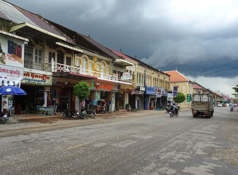 A long row of two-story shophouses in yellow and cream with shuttered upper windows and balconies above shop signs, along a wide street under a dark cloudy sky.