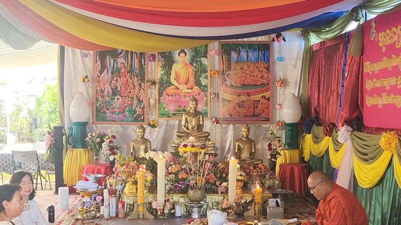 Altar under colourful draped fabric with three bronze seated Buddhas, candles, flowers and Buddha paintings, a monk seated at right.