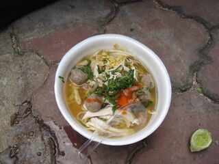 A white bowl of noodle soup with meatballs, sliced carrot and herbs, a lime wedge beside it.