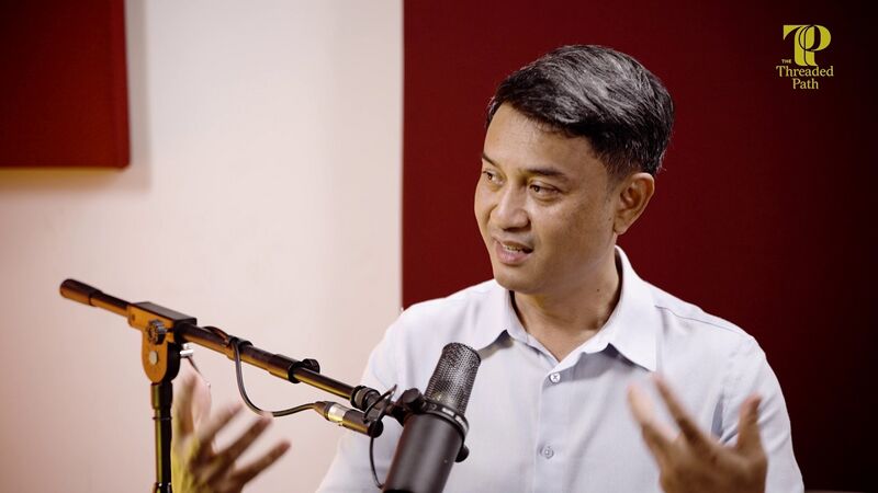 A man in a light blue shirt gestures with both hands as he speaks into a studio microphone against a dark red wall.
