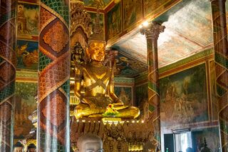 A golden seated Buddha statue in a hall whose walls and pillars are covered in colorful painted scenes.