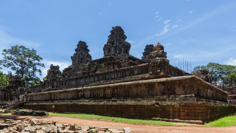 A stepped pyramid of dark, plain stone, seen from one corner, rises in tiers with long galleries to towers on its top terrace, under a blue sky. Loose stone blocks lie on the ground in front, a large tree stands at the left and scaffolding at the right edge.