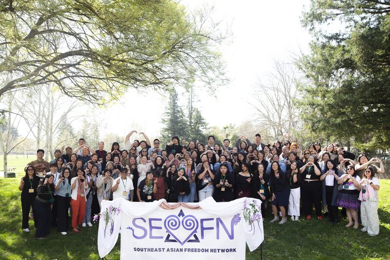 A large group of people in dark clothes stand on a lawn behind a white SEAFN banner, raising their hands.