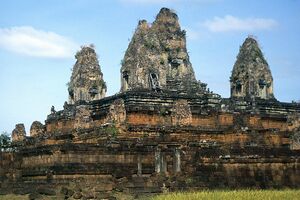 A stepped pyramid of weathered laterite and brick tiers rises to three crumbling brick towers with grass and small plants growing from their tops, under a blue sky with a few clouds. A wooden brace props one tower doorway, and dry grass fills the foreground.