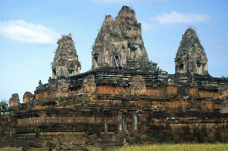A stepped pyramid of weathered laterite and brick tiers rises to three crumbling brick towers with grass and small plants growing from their tops, under a blue sky with a few clouds. A wooden brace props one tower doorway, and dry grass fills the foreground.