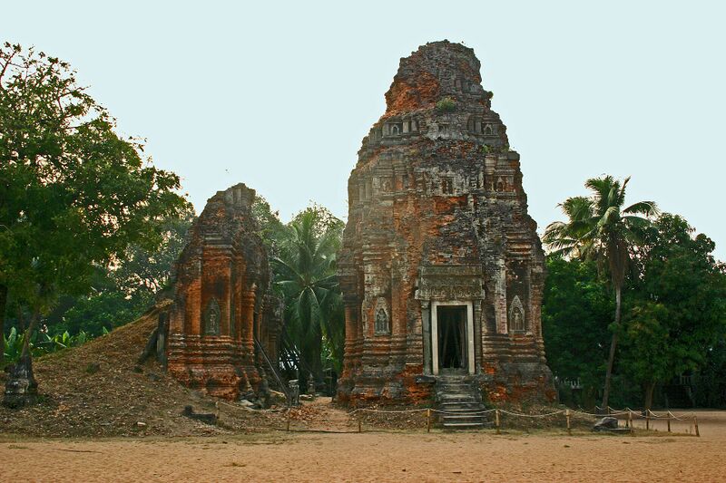 Two weathered brick temple towers, the taller with a carved doorway and steps, among palms and leafy trees, under a pale sky.