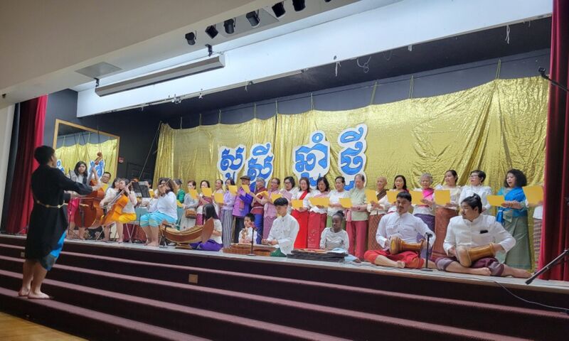 Singers and musicians in Cambodian dress on a stage under a gold banner reading Happy New Year in Khmer.