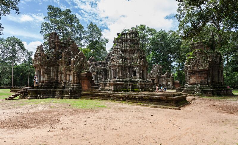 A small sandstone temple with a carved central tower, a long hall and a gateway, set on a low platform among tall trees.