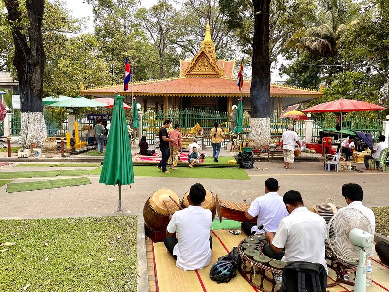 A small pavilion shrine with a tiered roof among trees, and musicians seated on mats in front