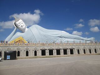 A giant white reclining Buddha statue on a long base under a blue sky.