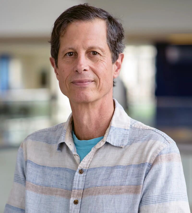 Man with short dark hair in a striped short-sleeved shirt, standing in a bright building.
